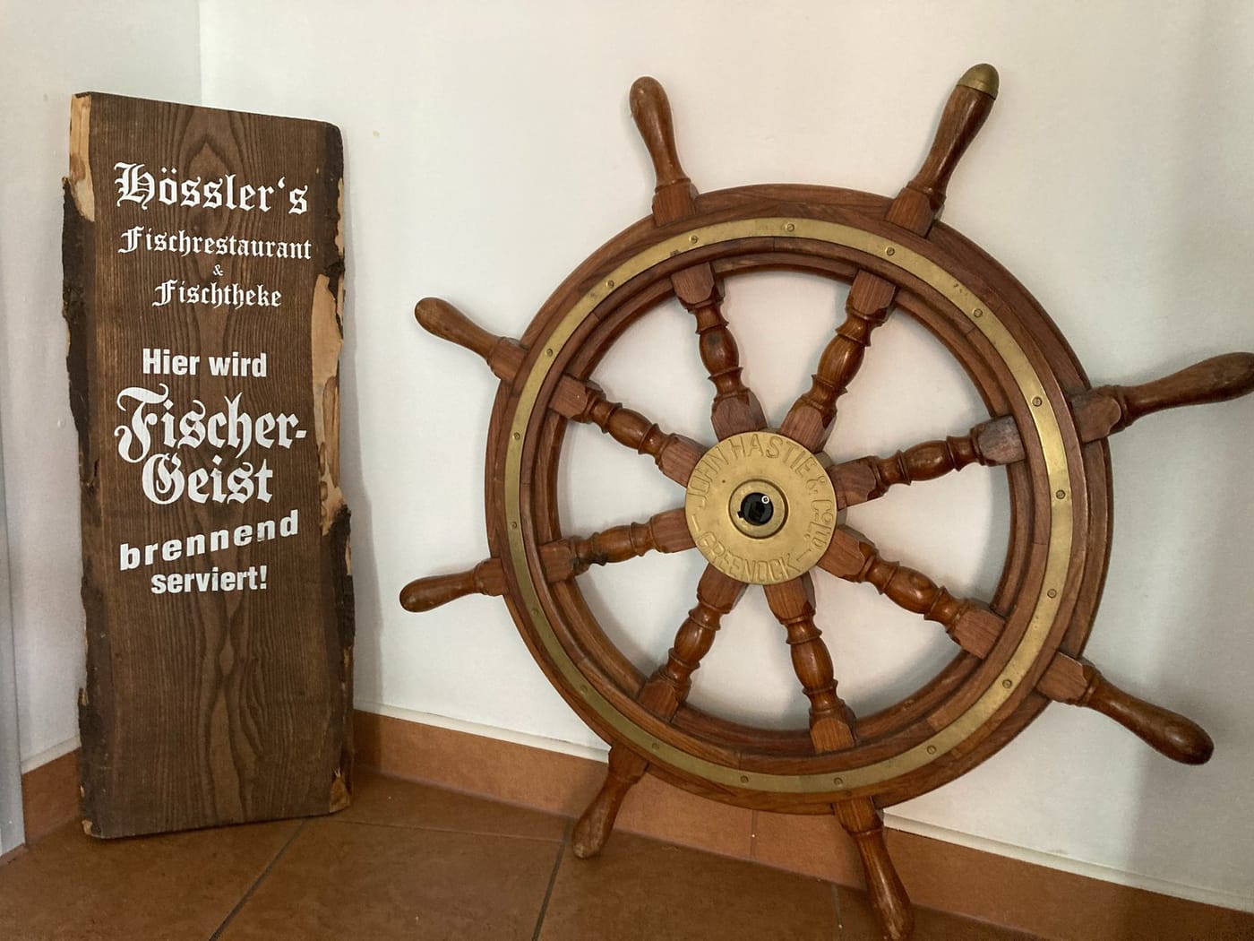 Holzschild „Hössler's Fischrestaurant & Fischtheke – Hier wird Fischer-Geist brennend serviert!“ neben einem Schiffsteuerrad